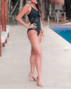 Woman in a black swimsuit standing by a poolside with palm trees in the background