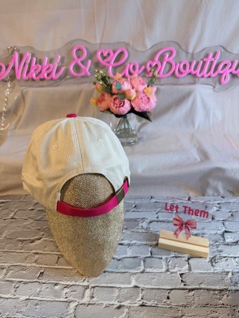 Beige cap with pink band on a mannequin head, floral arrangement, and 'Nikki & Co. Boutique' sign in the background.