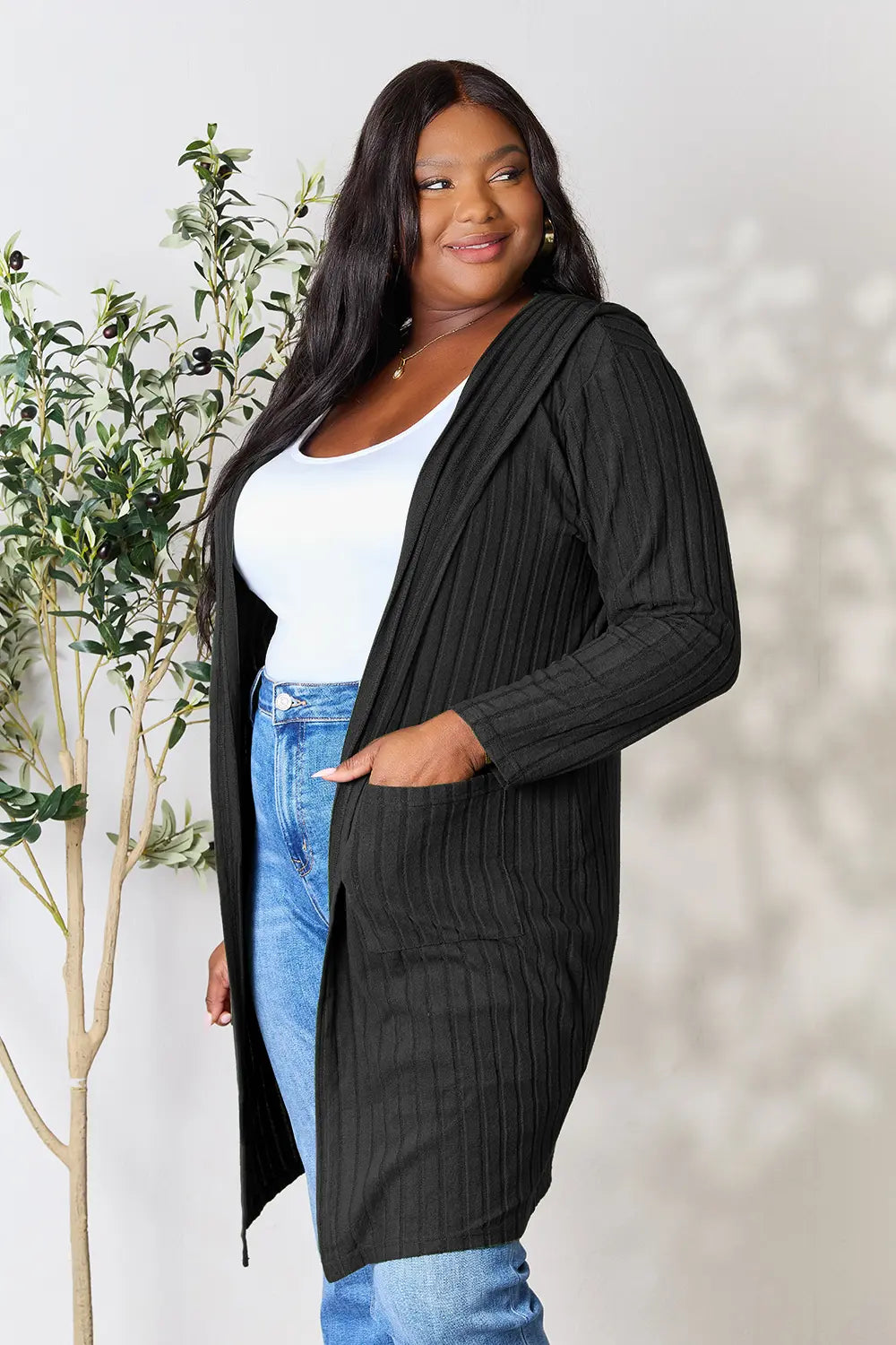 Woman wearing a black ribbed cardigan over a white top and blue jeans, standing next to a plant.