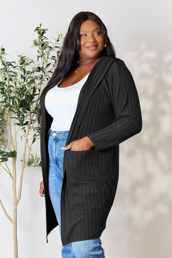 Woman wearing a black ribbed cardigan over a white top and blue jeans, standing next to a plant.