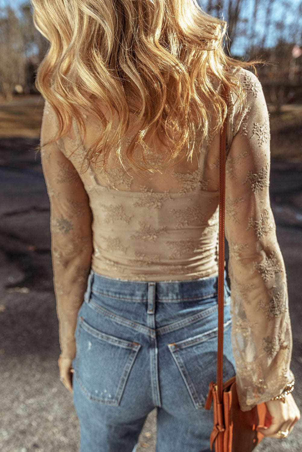 Person wearing a beige lace top and blue jeans with a brown leather bag outdoors.