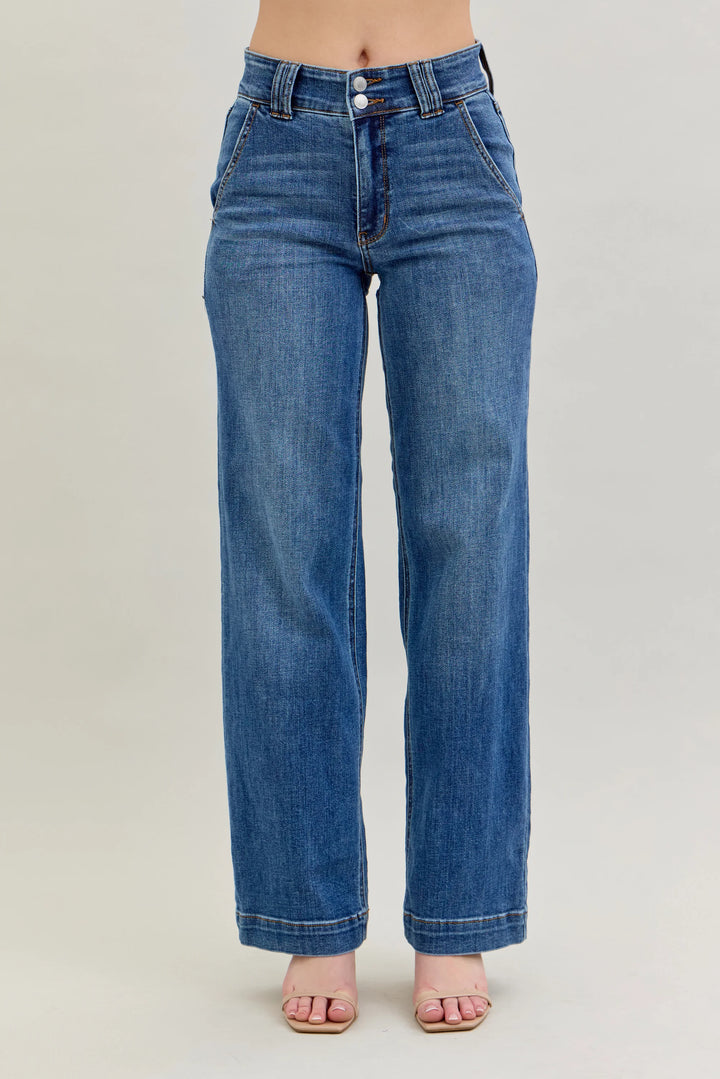 A person wearing medium-wash blue denim jeans with a straight-to-wide leg fit, shown from the waist down against a plain light gray studio background.