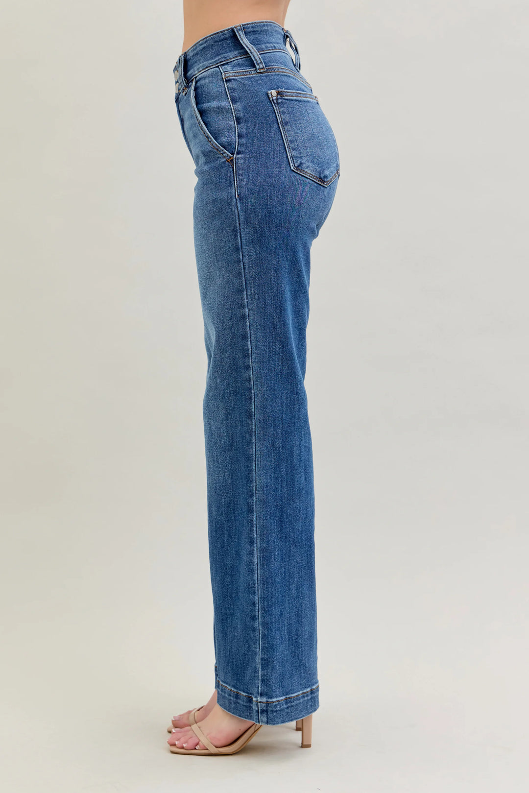 A side profile view of a person wearing blue high-waisted straight-leg jeans and light pink heeled sandals against a light gray studio background.