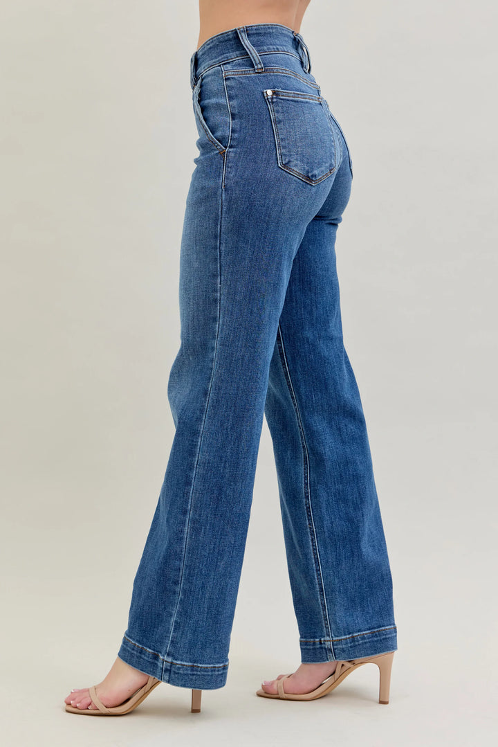 A person wearing blue high-waisted flare jeans with a side-back view, paired with nude heeled sandals, against a plain light beige studio background.