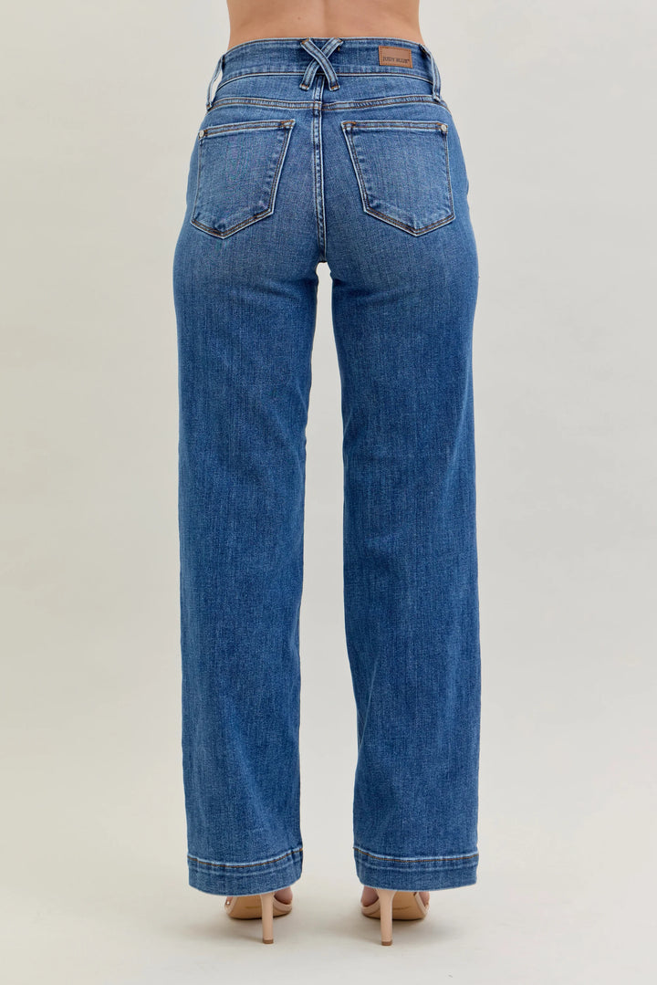 Rear view of a model wearing blue wide-leg denim jeans with a faded wash, paired with beige high heels, against a light gray studio background.