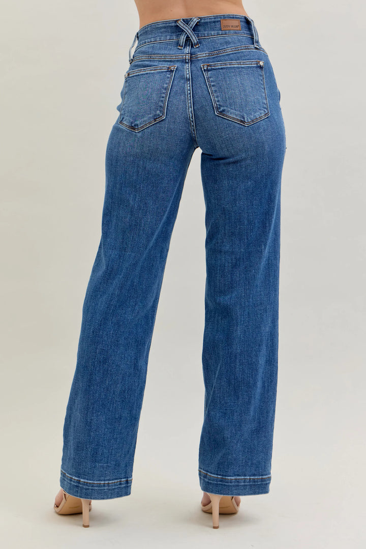 Rear view of a person wearing blue wide-leg denim jeans with beige high-heeled sandals against a light gray studio background.