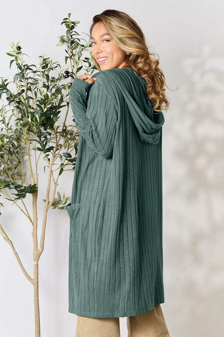 Woman wearing a green ribbed cardigan standing next to a plant against a white background