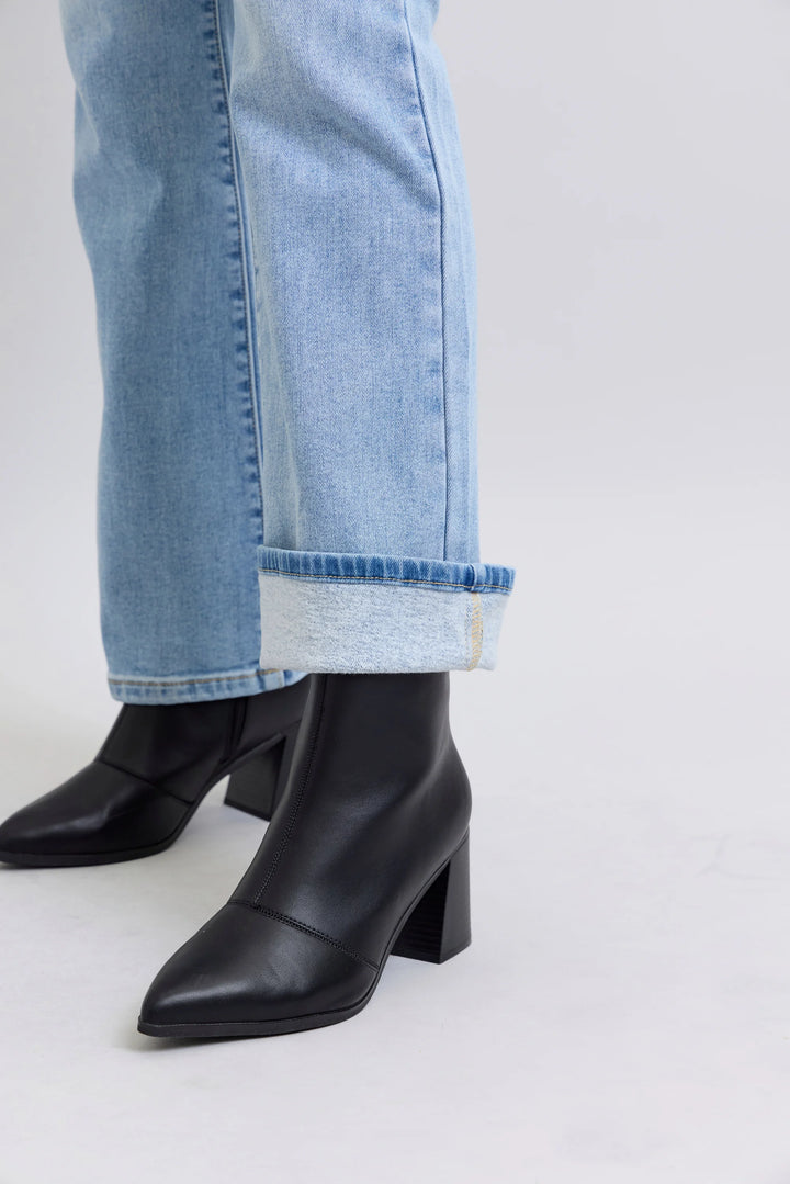 Black leather ankle boots with a block heel and pointed toe, worn with light blue cuffed jeans against a light gray studio background.