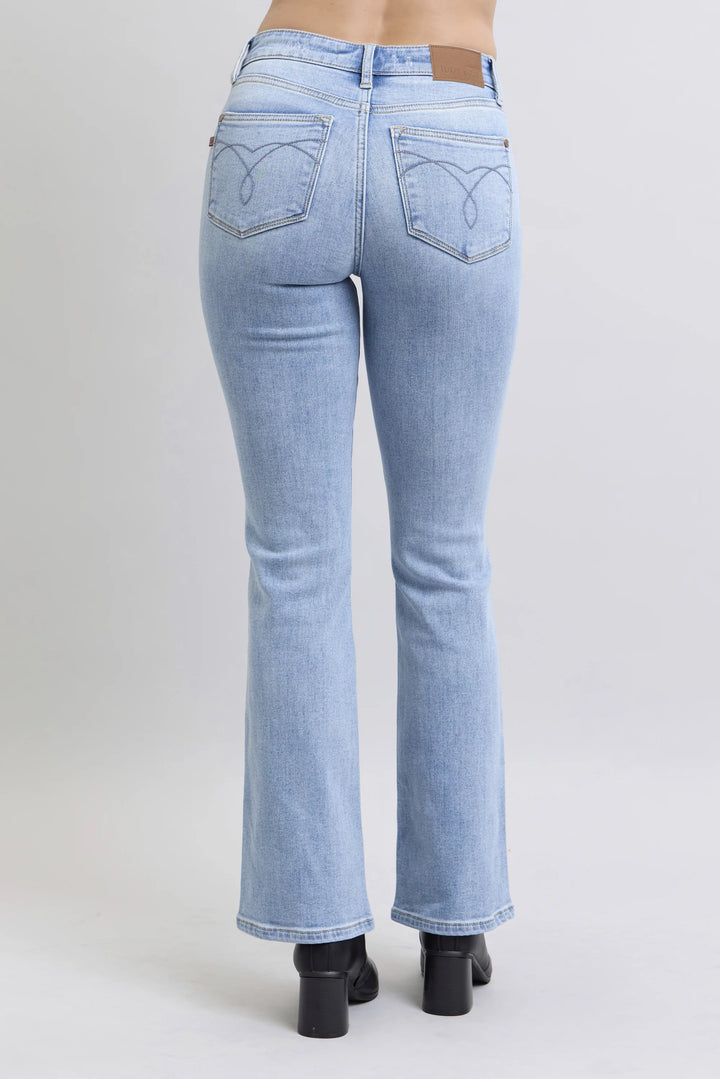 Rear view of a person wearing light-wash blue flare jeans with black block heels, shown against a plain light gray studio background.