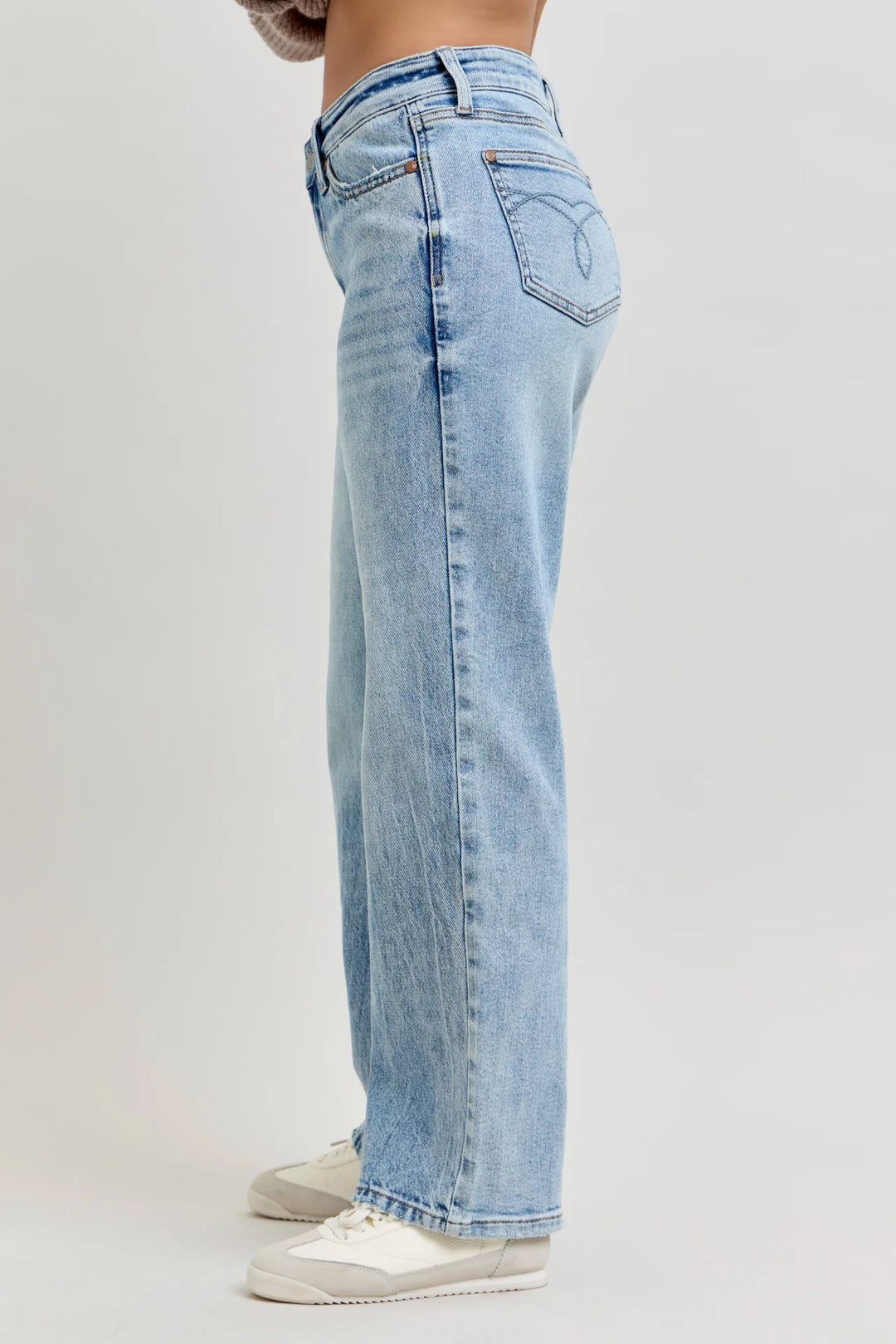 A side profile view of a person wearing light-wash blue denim jeans and white sneakers, standing against a plain light gray studio background.