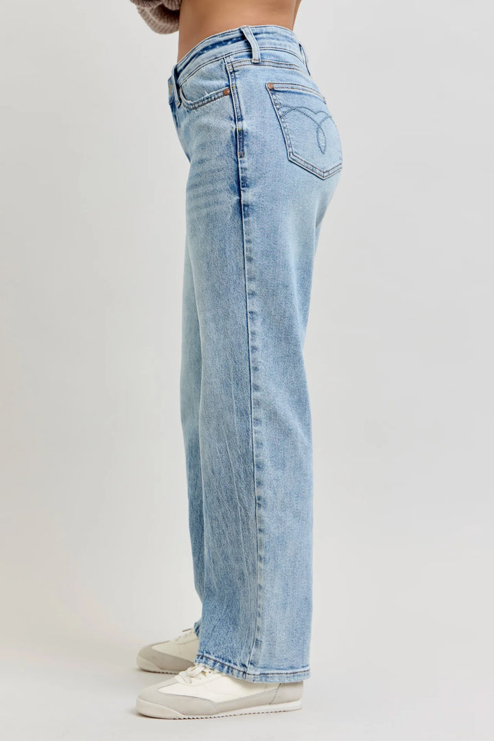 A side profile view of a person wearing light-wash blue denim jeans and white sneakers, standing against a plain light gray studio background.