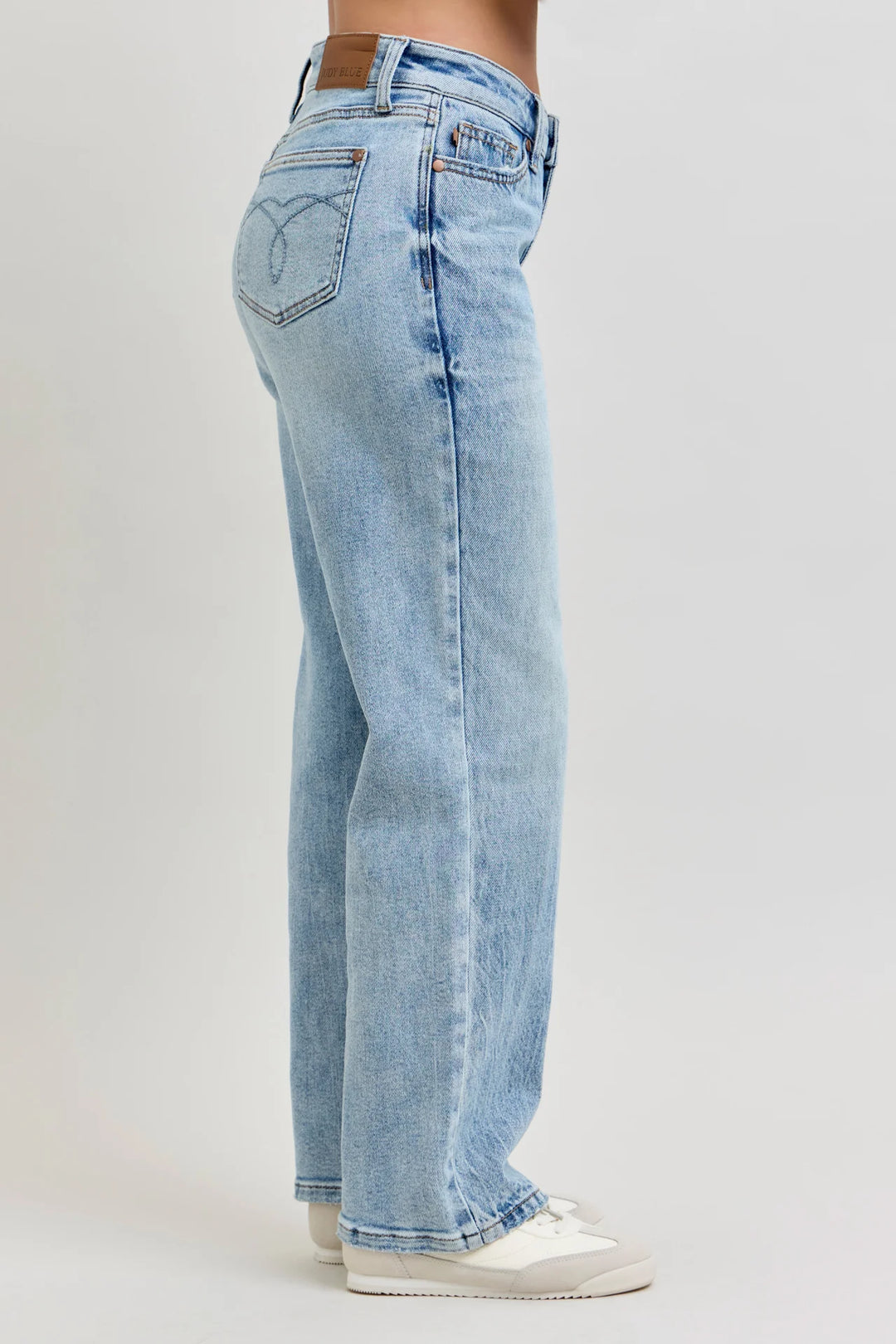 A side profile view of a person wearing light-wash blue denim jeans and white sneakers against a plain light gray studio background.