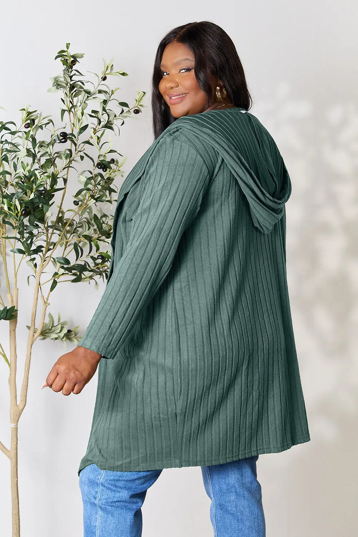 Woman wearing a green ribbed sweater standing next to a plant against a white wall