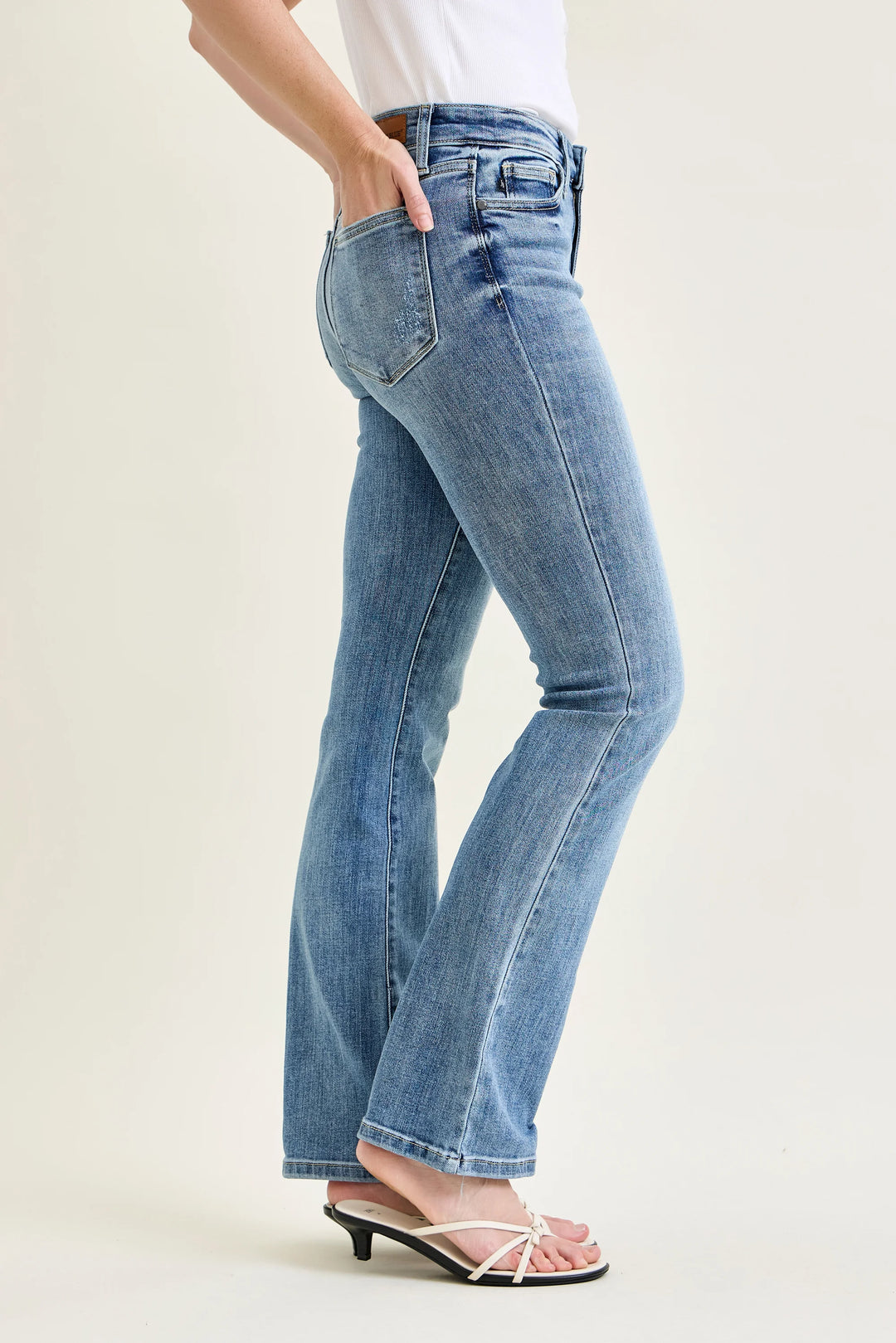 A person wearing light blue high-rise flare jeans with a side pocket, paired with a white top and white heeled thong sandals, shown from the waist down against a plain light beige studio background.