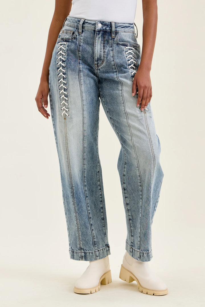 A model wearing light-wash blue jeans with white lace-up detailing on the front of both legs, paired with white ankle boots, against a plain off-white studio background.
