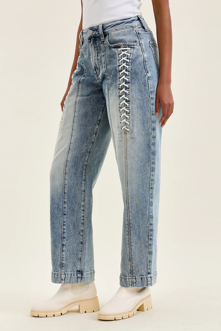 A model wearing light-wash blue jeans with decorative white lace-up detailing down the side of the right leg, paired with white block-heel boots against a plain light beige studio background.