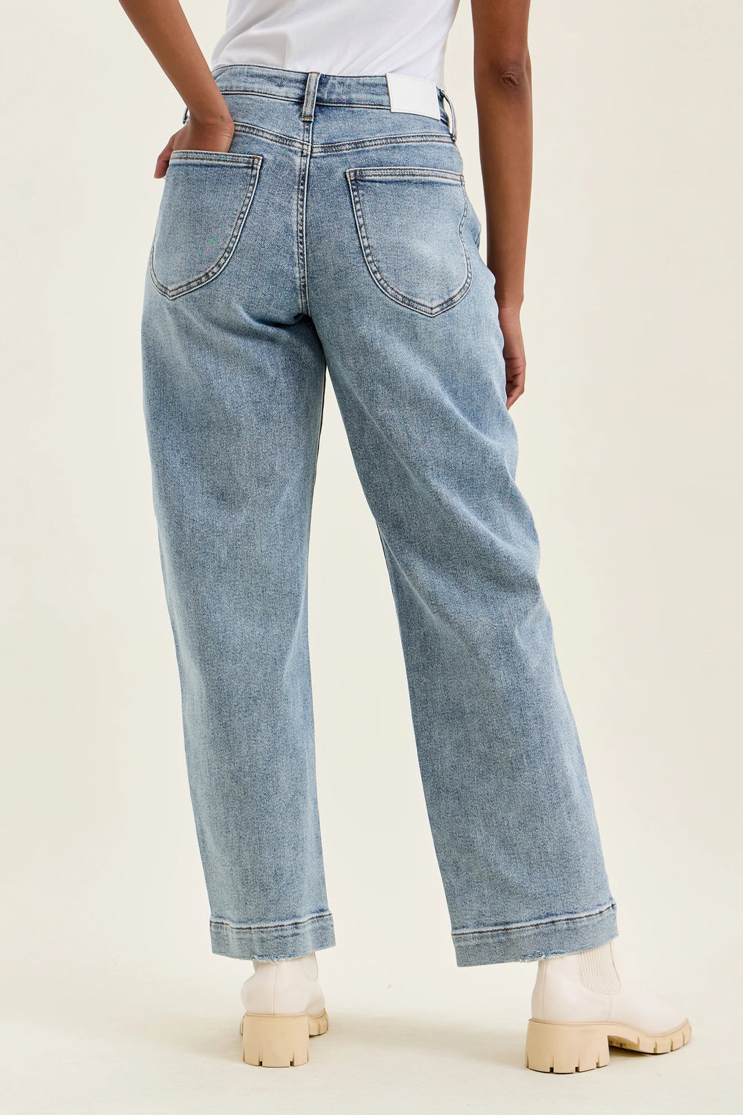 A person shown from the back wearing light-wash blue jeans with a straight-leg or wide-leg fit, paired with white socks and beige block-heel shoes against a plain light beige studio background.