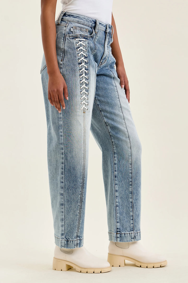 A person wearing light-wash blue jeans with decorative white lace-up detailing down the front of the right leg, paired with off-white ankle boots against a plain light beige studio background.
