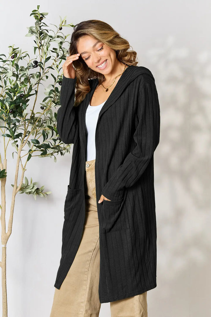 Woman wearing a black cardigan over a white top and beige pants, standing against a light background with a plant to the left.