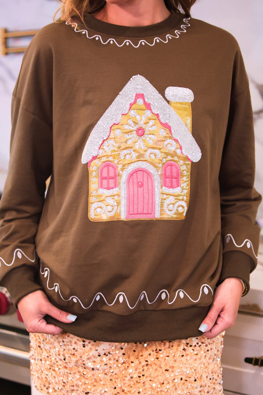 A person wearing a brown long-sleeve sweater with a gingerbread house applique on the front, white wavy trim around the neckline and hem, and a sparkly beige skirt.
