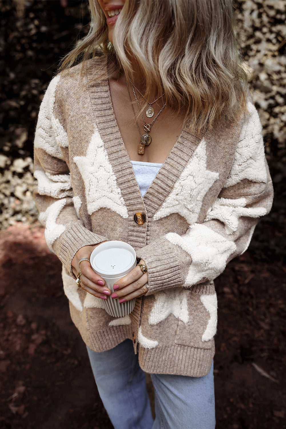 Person wearing a beige cardigan with star patterns, holding a white mug outdoors.
