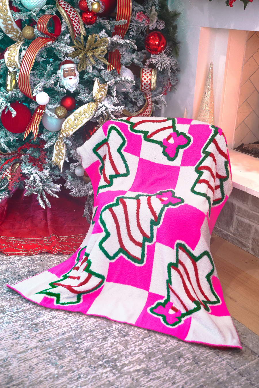 A bright pink and white checkered blanket with green-outlined Christmas tree patterns draped over the front of a decorated Christmas tree in a living room setting.