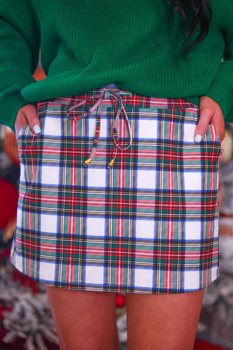 A close-up of a person wearing a green knit sweater and a white plaid mini skirt with red, green, and blue stripes, holding the sides of the skirt to show its shape.