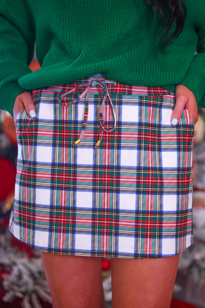 A close-up of a person wearing a green knit sweater and a white plaid mini skirt with red, green, and blue stripes, holding the sides of the skirt to show its shape.