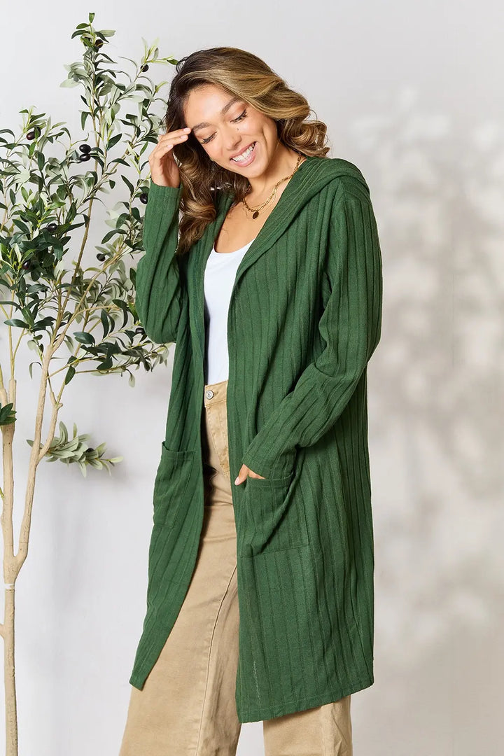 Woman wearing a green cardigan standing next to a plant against a white background