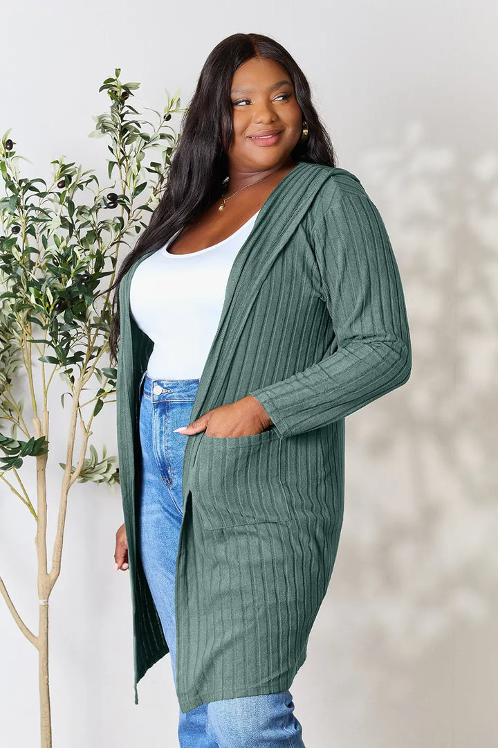 Woman wearing a green ribbed cardigan over a white top and blue jeans, standing next to a plant.
