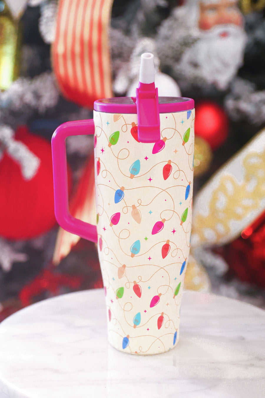 A tall insulated travel mug with a handle and straw lid, decorated with a Christmas light pattern on a cream background, with bright pink accents on the handle, lid, and straw. The mug is set on a white marble surface with a blurred festive holiday tree background.