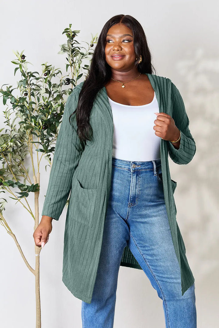 Woman wearing a green cardigan, white top, and blue jeans standing next to a plant.