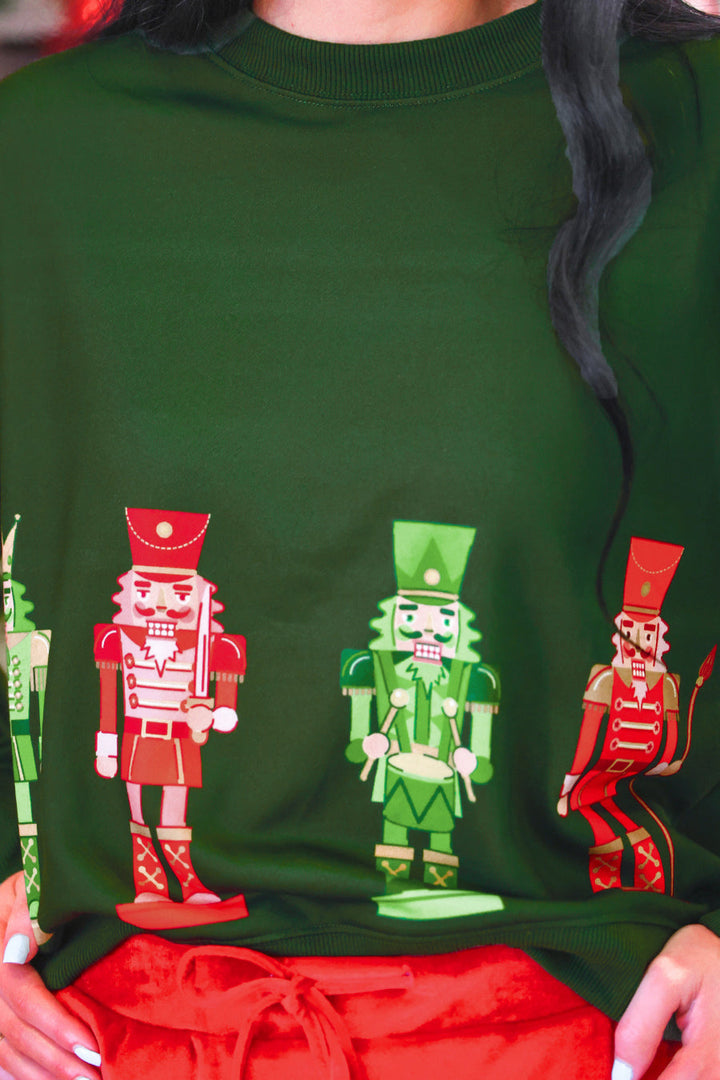 A close-up of a person wearing a dark green crewneck sweatshirt with a graphic print of three nutcracker figures in red and green, paired with red pants.