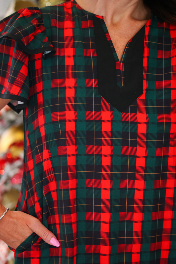 A close-up of a woman wearing a festive holiday plaid top with red, green, and black tartan checkered pattern, featuring a black V-neckline panel.