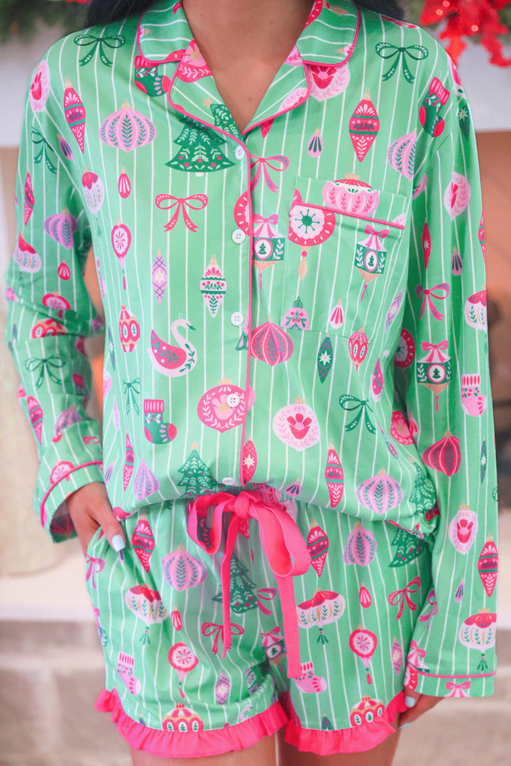 A person wearing a matching holiday-themed pajama set with a green and white striped long-sleeve top and shorts featuring pink trim and festive motifs like ornaments, bows, trees, and stockings.