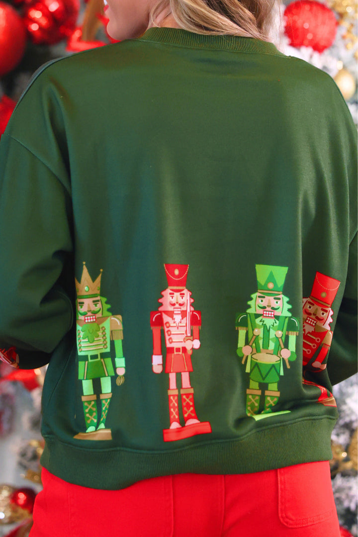 A person viewed from behind wearing a dark green crewneck sweatshirt with a graphic of four colorful nutcracker figures across the back, paired with bright red pants. The background is a festive holiday setting with red ornaments and lights.