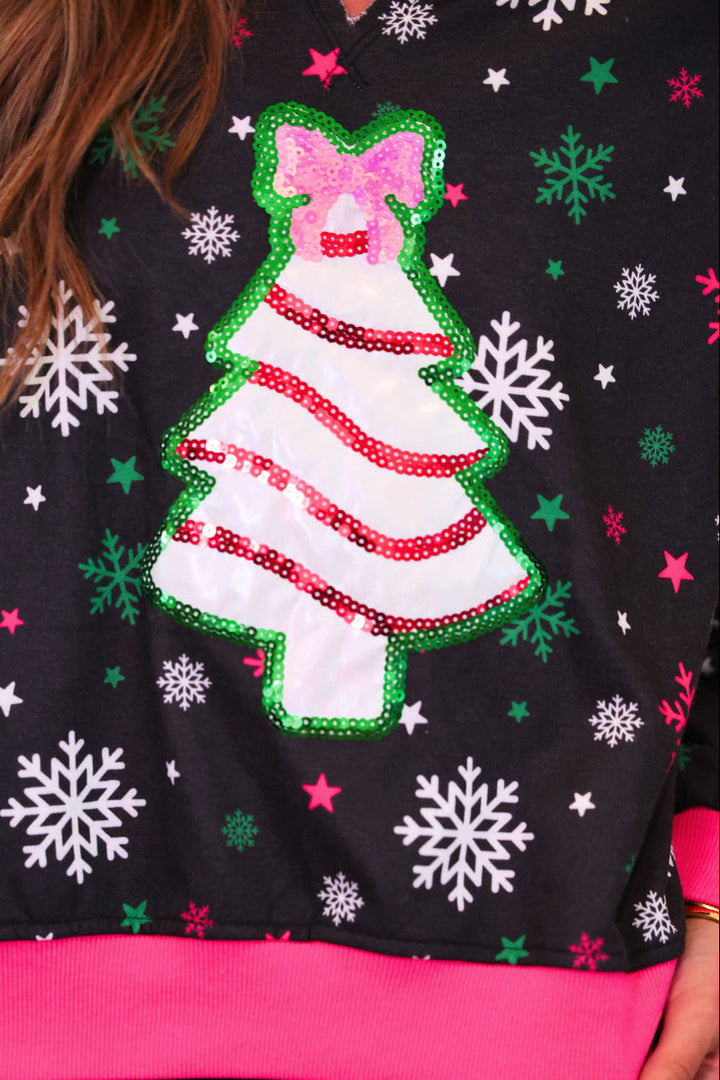 A close-up of a black Christmas-themed top featuring a sequined white Christmas tree with a pink bow, surrounded by white and green snowflakes and stars, worn by a person.