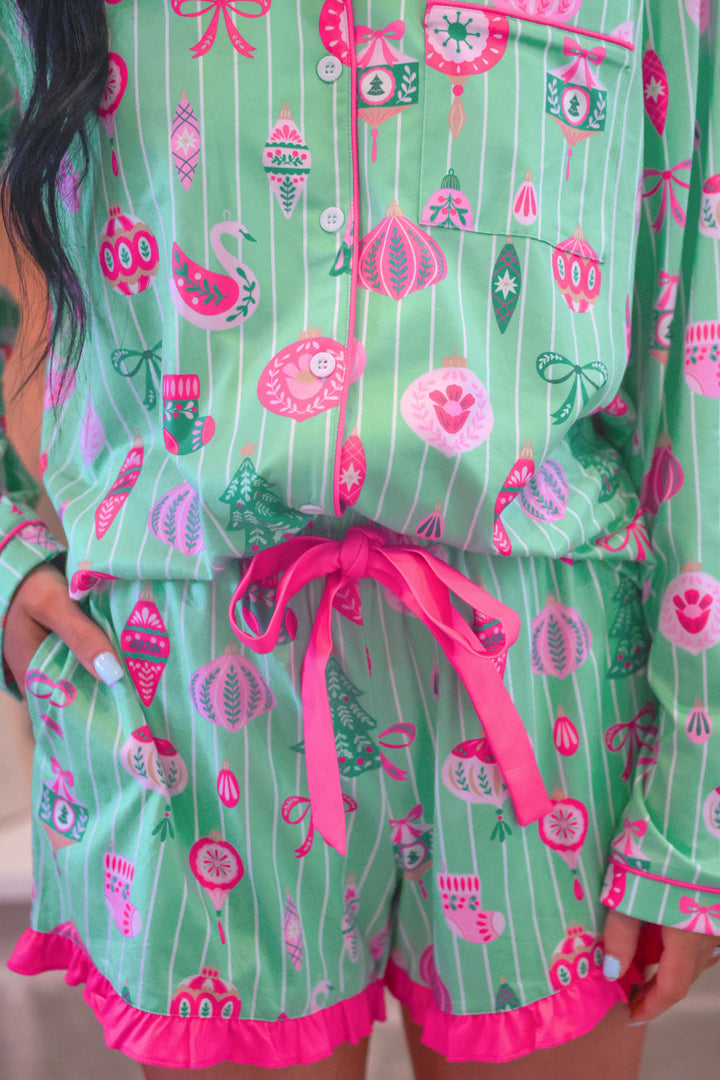 A close-up of a person wearing a matching pajama set with a festive holiday print. The set features a light green base with thin white vertical stripes and colorful motifs of ornaments, snow globes, Christmas trees, and houses in pink, red, and dark green.