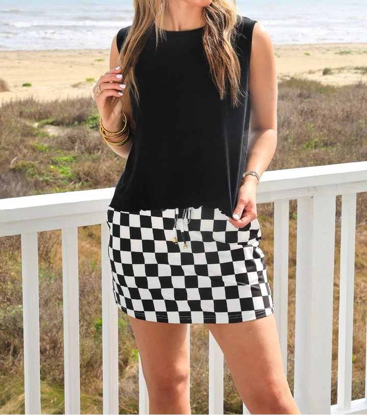Woman wearing a black top and checkered skirt standing on a balcony with a beach in the background