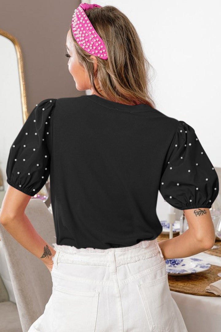 Woman wearing a black top with white pearl dots and white pants, standing in a room.