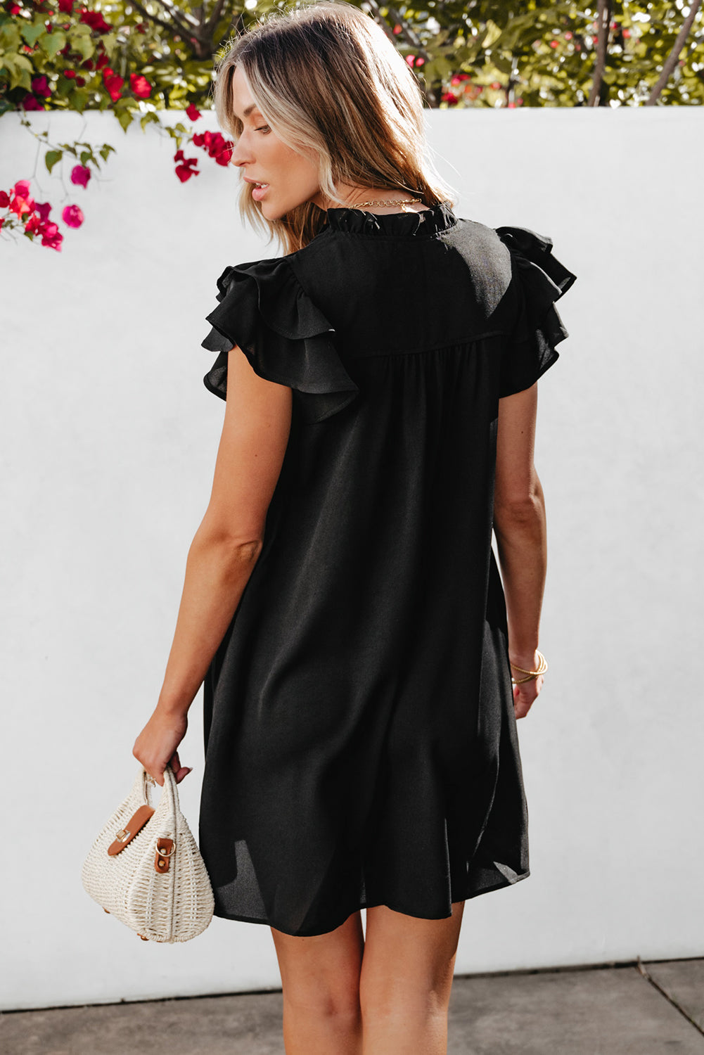 Woman wearing a black dress with ruffled shoulders against a white wall with greenery.