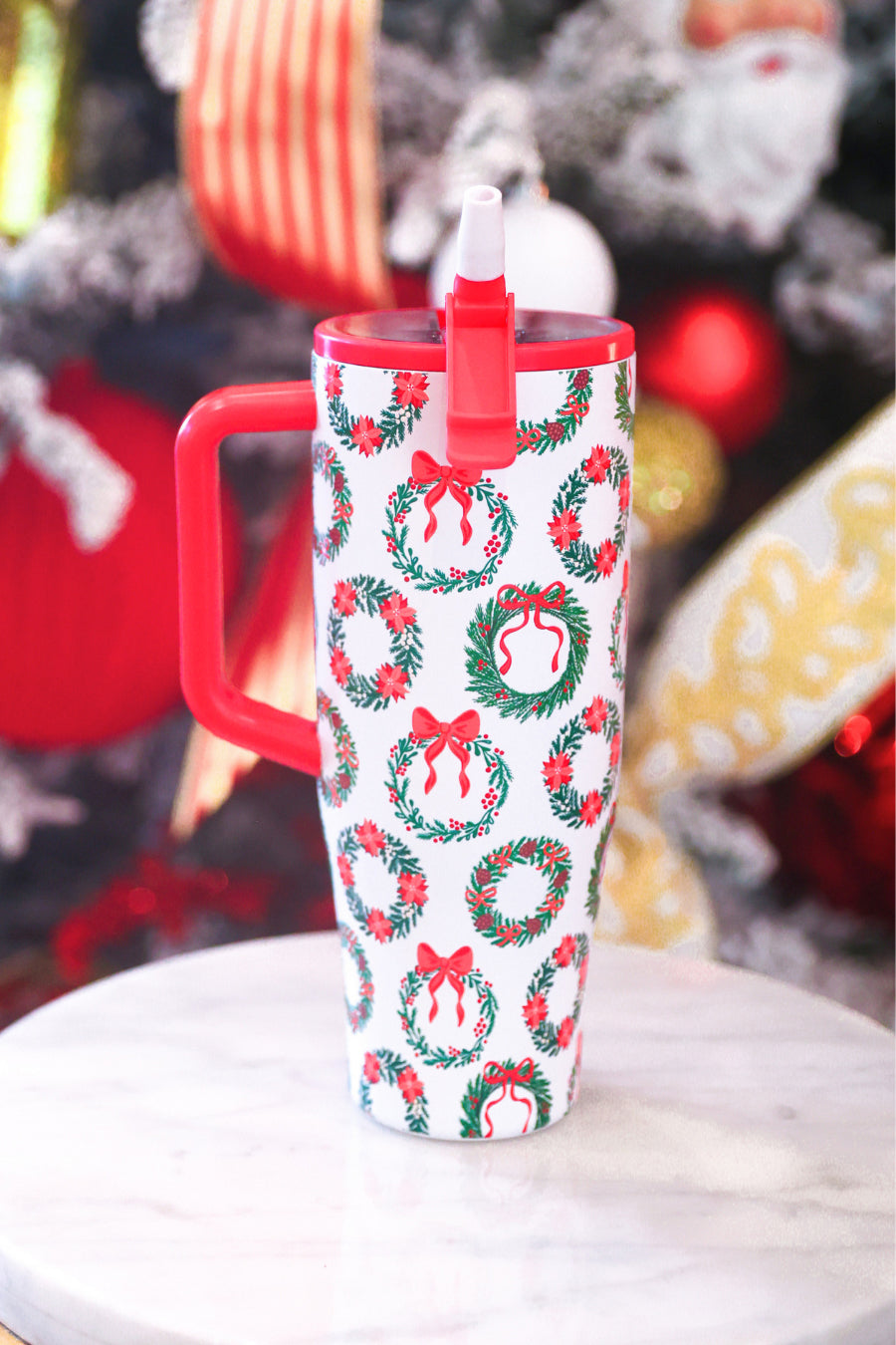 A tall insulated travel mug with a red handle and lid, featuring a white body printed with green wreaths and red bows, set on a light marble surface with a blurred Christmas tree in the background.