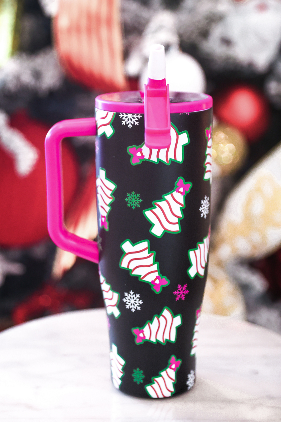 A black insulated tumbler with a bright pink handle and lid, decorated with a festive Christmas tree pattern in white, green, and pink. The tumbler is shown on a light marble surface with a blurred holiday background.