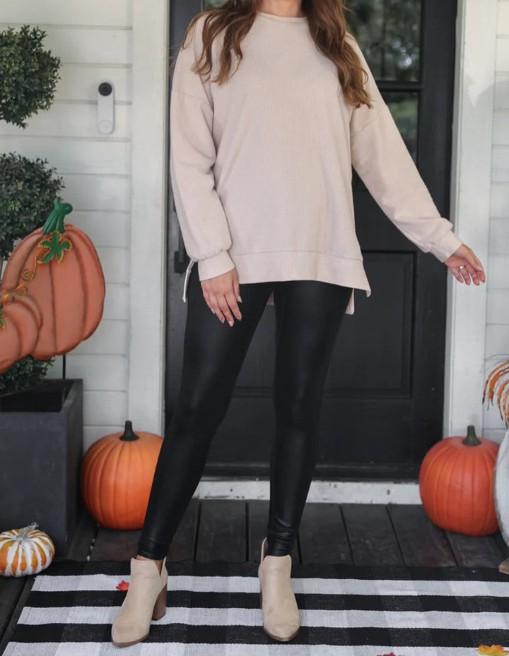 A woman stands on a porch wearing a light beige crewneck sweatshirt, black leggings, and beige ankle boots. The porch has a black door, white siding, pumpkins, and a black-and-white striped rug.