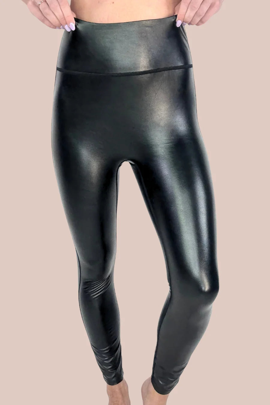 High-waisted black faux-leather leggings with a glossy finish, shown on a model against a solid beige background.