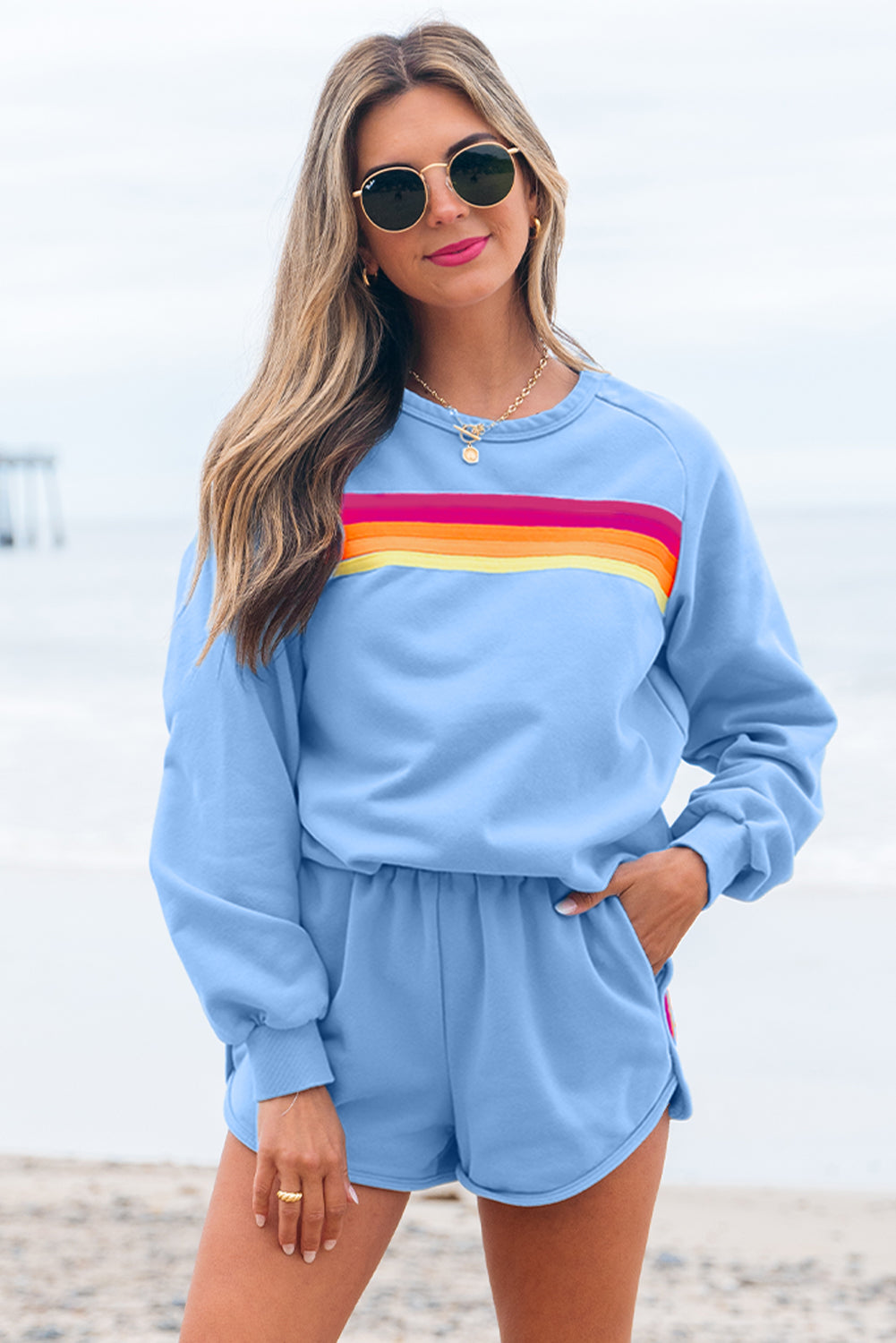 Woman wearing a light blue sweatshirt with rainbow stripes and matching shorts on a beach.