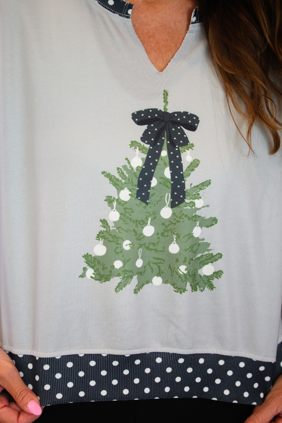 A close-up of a person wearing a light gray V-neck top with a green Christmas tree graphic on the front, decorated with white ornaments and a dark navy polka-dot bow. The bottom hem of the top features a matching dark navy and white polka-dot band.
