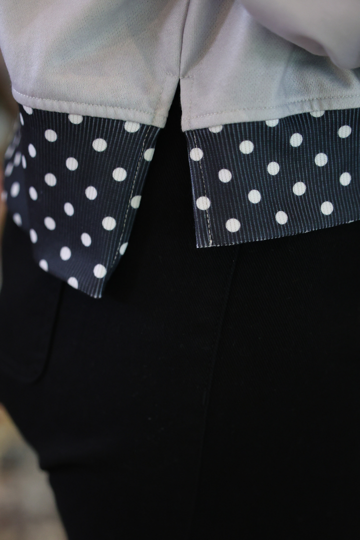 Close-up of a garment with a light gray upper section and black lower section, featuring contrasting black-and-white polka dot ribbon trim at the waist or pocket opening.