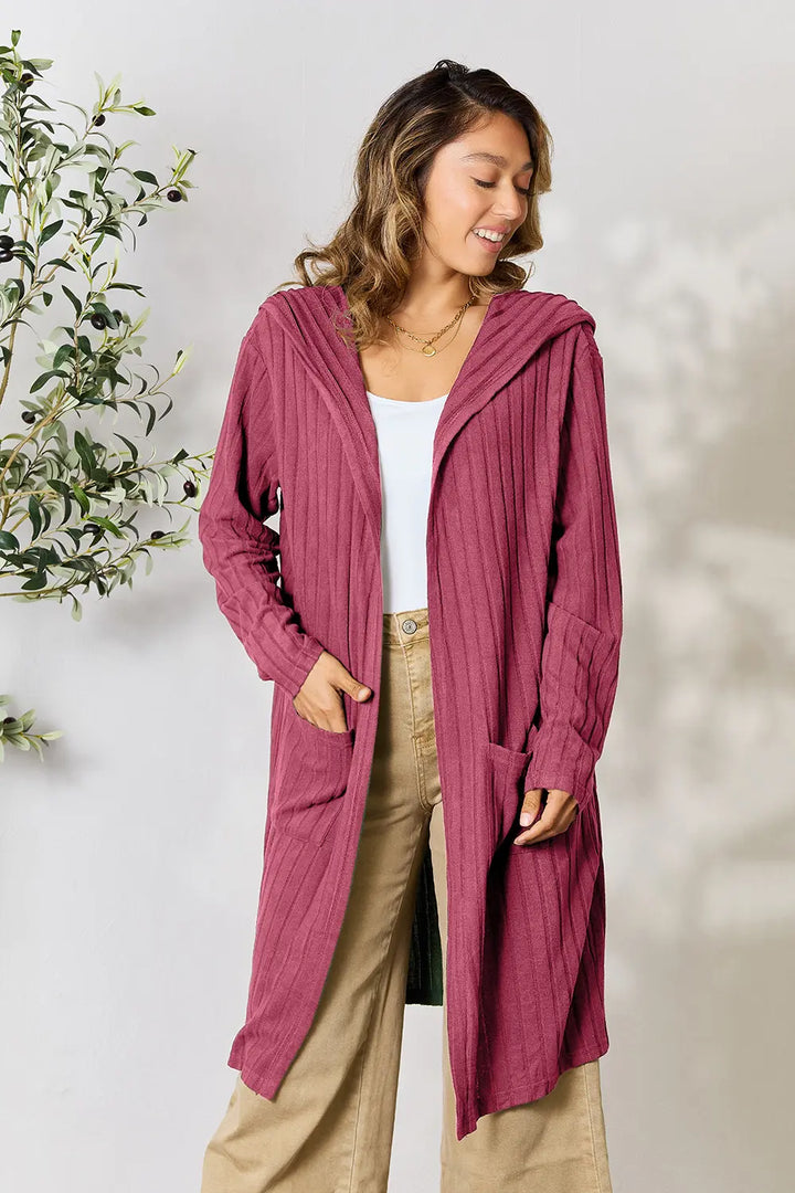 Woman wearing a long pink cardigan with a white top and beige pants, standing against a light background.
