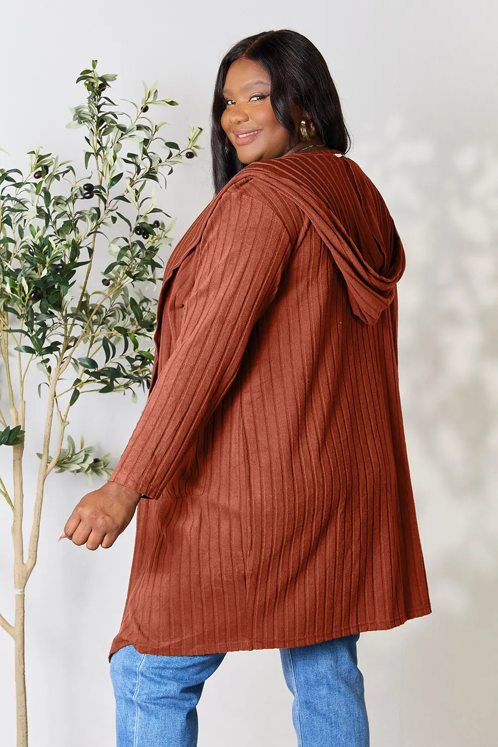 Woman wearing a rust ribbed sweater and blue jeans against a white background with a plant.